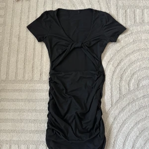 Svart kort klänning med knyt - Svart kort klänning med knyt framtill och cut-out under bysten. Klänningen har korta ärmar, djup urringning och rynkade sidor som ger en snygg passform. Perfekt för dig som gillar trendiga detaljer och en stilren look.