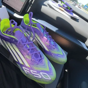 Adidas F50 lila fotbollsskor - Säljer ett par Adidas F50 fotbollsskor i en riktigt fet lila färg med neon-gula och vita detaljer. Skorna har snörning och en aerodynamisk design med tydliga F50- och Adidas-loggor på sidorna. Perfekta för dig som vill sticka ut på planen. Det är ett par F50 AG Elite. Nypris 3000kr, mitt pris 1400kr. Skriv gärna vid frågor
