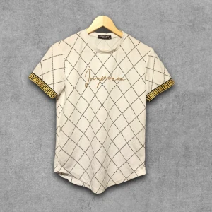 Beige rutig t-shirt från Johnny Voga - Snygg beige t-shirt från Johnny Voga med svart rutmönster och gulddetaljer på ärmsluten. T-shirten har korta ärmar och ett broderat guldigt signaturtryck på bröstet. Perfekt för dig som gillar unika detaljer och stilren design.