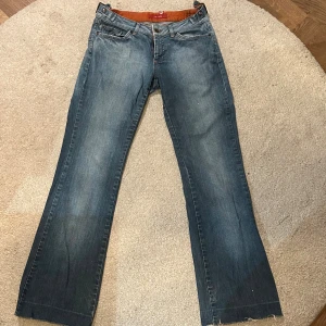 Blå jeans med bootcut och orange detaljer - Snygga blå jeans från VM Jeans med bootcut passform och klassiska fem fickor. Jeansen har orange detaljer på insidan av linningen och kontrastsömmar på bakfickorna. Tillverkade i denim med slitna detaljer nertill för en avslappnad look. De är slitna nertill (ses på bild 2) men annars inga tydliga skador. De är ca 37 cm i midjan och ca 100cm från midjan nertill och själva benet är ca 73cm. Säjer då de tyvärr inte passar mig.
