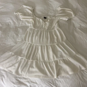 Vit somrig kortklänning med puffärm - Superfin vit kortklänning med puffiga kortärmar och volanger i flera lager 💕. Klänningen har en söt snörning framtill och är tillverkad i ett lätt, strukturerat tyg som känns luftigt och fräscht.