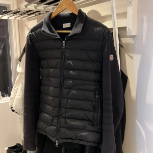 Moncler Cardigan - Snygg svart pufferjacka från Moncler med quiltad front och mjuka stickade ärmar. Jackan har dragkedja framtill, ståkrage och Moncler-logga på ärmen. Perfekt för dig som vill ha en stilren och sportig look. Säljer pågrund av att ha tröttnad på den men annars inga defekter eller fel!
