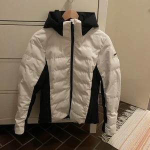 Vit och svart jacka från Everest - Säljer en snygg pufferjacka från Everest i storlek 38. Jackan är vit med svart detalj på sidan. Den har dragkedja framtill, två fickor och en mysig huva. Perfekt för kalla dagar när du vill hålla dig varm och se cool ut.