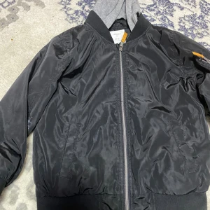 Svart bomberjacka från Lindex Kids - Svart bomberjacka från Lindex Kids med klassisk ribbad krage och muddar. Jackan har dragkedja framtill, två fickor och en ärmficka med orange detalj. Vattenavvisande material, perfekt för höst och vår. Enkel och stilren design som passar till det mesta.