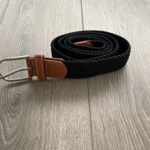 Svart flätat bälte med läderdetaljer - Snyggt svart flätat bälte med bruna läderdetaljer och silverfärgat spänne. Bältet har en stilren design och passar perfekt till jeans eller chinos. Flätningen gör det flexibelt och lätt att justera efter önskad längd.