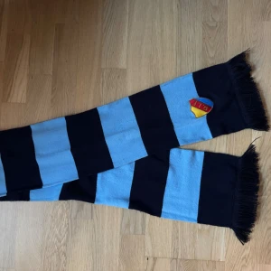 Randig Djurgården halsduk blå/blå - Lång halsduk med breda ljusblå och marinblå ränder samt fransar i ändarna. Broderad Djurgården-logga i ena änden. 