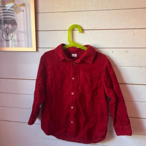 Röd manchesterskjorta med knappar - Snygg röd skjorta i manchester med klassisk krage och bröstficka. Skjortan har långa ärmar och stängs med ljusa knappar framtill. Perfekt för dig som vill ha en färgstark och trendig look.