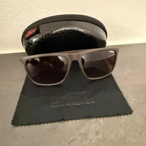Bruna Carrera solglasögon med etui - Snygga solglasögon från Carrera i matt brun plast med fyrkantig form och mörka linser. Sidorna har en diskret textur och Carrera-logga. Kommer med svart hårt etui med ormskinnsmönster och putsduk. Perfekta för att lyfta din stil i solen.
