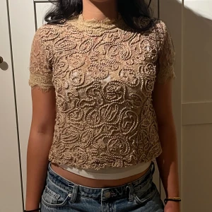 Beige spetsblus med broderi - Säljer en beige, kortärmad blus med vackra broderade detaljer och spets vid ärmslut och hals. Blusen har en croppad passform och ett unikt, mönstrat tyg som ger en lyxig känsla. Perfekt för dig som vill sticka ut med en elegant och trendig topp.Dragkedja där bak som går att öppna hela vägen. 