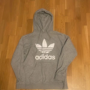 Grå Adidas hoodie med stor logga - Säljer en grå hoodie från Adidas med stor vit trefoil-logga och text på bröstet. Tröjan har huva med snörning och en klassisk känguruficka framtill. Perfekt för en chill och sportig stil.