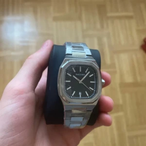 Faucon silverfärgad klocka - Stilren klocka från Faucon med svart urtavla och datumvisning. Boetten är åttakantig och armbandet är i silverfärgad metall med snygg länkdesign. Klockan har minimalistiska index och klassisk krona på sidan. Perfekt accessoar för en clean look.