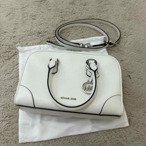 Michael Kors Handväska - Helt Ny - Stilren och elegant handväska från Michael Kors i vitt med klassisk MK-detalj framtill. Väskan är helt ny och oanvänd.  Detaljer: • Färg: Vit med svarta kanter • Material: Slitstarkt konstläder • Justerbar axelrem + handtag • Flera fack inuti för nycklar, mobil och små tillbehör • Snygg och kompakt design – perfekt för både vardag och fest  En lyxig, mindre väska i hög kvalitet som ger en exklusiv och modern look.