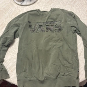 Olivgrön sweatshirt från Vans - Olivgrön sweatshirt från Vans med stort logotryck i camo-stil på bröstet. Tröjan har rund hals, ribbade muddar och är tillverkad i mjuk bomullsmix. Perfekt för en avslappnad streetwear-look. Den är storlek small men passar medium också enligt mig