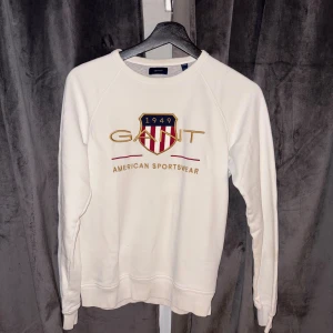 Vit sweatshirt från Gant med brodyr - Vit sweatshirt från Gant med broderad logga och sköld på bröstet. Klassisk rund halsringning och långa ärmar. Snygg och stilren tröja i mjuk bomull, perfekt för en sportig och avslappnad look.