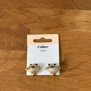 Två par clipsörhängen  - Säljer två par clipsörhängen. Ett par är guldiga med solfjäderform och gnistrande stenar, det andra paret är rundade och blanka i plast med guldfärg. Båda har klassisk retrostil och är perfekta för dig som vill ha statementörhängen utan hål i öronen. Obs de är inte från glitter, använde bara förpackningen för att ta bilder 