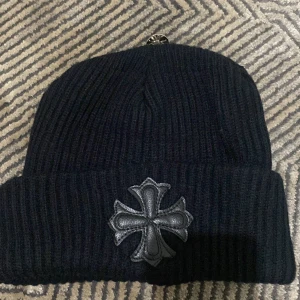 Svart ribbad mössa Chrome Hearts - Svart ribbstickad mössa från Chrome Hearts med ikonisk silverfärgad knapp och ett stort svart läderpatch i form av ett kors framtill. Mössan har uppvikt kant och är riktigt snygg för dig som gillar streetwear med exklusiva detaljer.