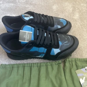 Valentino Garavani Rockrunner sneakers - Snygga Valentino Rockrunner sneakers med svart och blått kamouflagemönster. Skorna har svarta snören, platt sula och detaljer i mocka och skinn. Ikonisk logga på tungan och coola nitar på hälen. Perfekt för dig som gillar streetstyle och exklusiva märken.