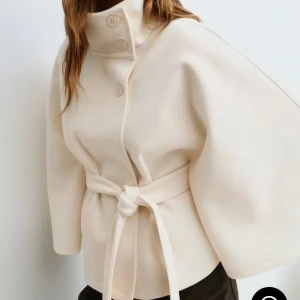 Beige kappa med bälte och hög krage - Stilren beige poncho kapp med hög krage, knappar framtill och knytskärp i midjan. Aldrig använt😊