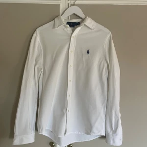 Vit långärmad skjorta från Ralph Lauren - Snygg vit skjorta från Ralph Lauren i featherweight mesh med klassisk krage och knappar framtill. Långärmad modell med det ikoniska blå Ralph Lauren-logot broderat på bröstet. Perfekt för dig som gillar stilrena och tidlösa plagg.