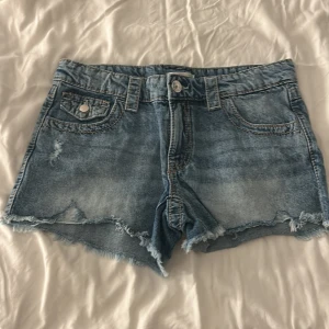 Blå jeansshorts från Gina Tricot - Snygga blå jeansshorts från Gina Tricot med fransig kant och slitna detaljer. Klassisk femficksmodell med knappar och bälteshällor. Perfekta för varma dagar och har en avslappnad, trendig look.