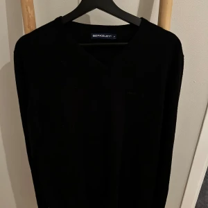 Svart ulltröja från Berkeley *L* - Klassisk svart långärmad tröja från Berkeley i fin ull. Tröjan har v-ringning och diskret broderad logga på bröstet. Perfekt för dig som gillar stilrena och enkla plagg med en snygg passform.