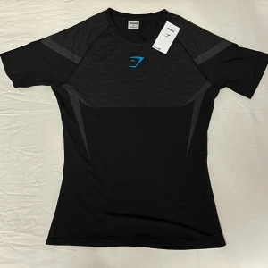 Gymshark Onyx - Svart t-shirt från Gymshark med korta ärmar och sportig passform. Framsidan har ett diskret mönster och en blå Gymshark-logga på bröstet. Materialet är lätt och stretchigt, perfekt för träning. Ett till ett. Snygga grå detaljer på axlar och rygg för extra stil. Denna tröja är limited edition, alltså går det inte att få tag på. Vid någora funderingar är det att bara att fråga 🤗. 