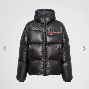 Svart pufferjacka från Prada - Säljer en svart pufferjacka från Prada med glansig finish och ikonisk röd logga på bröstet. Jackan har huva, dragkedja framtill och två fickor med dragkedja. Perfekt för kalla dagar och ger en riktigt clean och modern look. Storlek L nyskick 