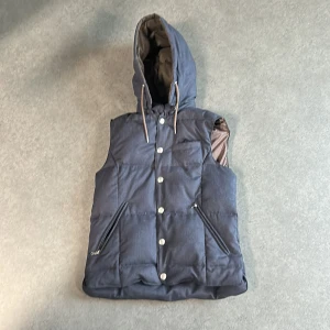 Mörkblå dunväst från Hansen & Jacob - Snygg mörkblå dunväst från Hansen & Jacob med huva och dragsko. Västen har knappar framtill, två dragkedjefickor och ett coolt paisleymönstrat innerfoder. Perfekt för lager-på-lager och chillare dagar.