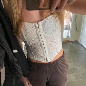 Vit broderad korsetttopp med spetsdetaljer - Vit korsetttopp med broderade spetsdetaljer och hålmönster. Aldrig använd