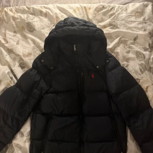 Svart pufferjacka från Polo Ralph Lauren - Svart pufferjacka från Polo Ralph Lauren med huva och dragkedja framtill. Jackan har två sidofickor med dragkedja och klassisk röd Polo-logga på bröstet. Perfekt för kalla dagar och riktigt snygg streetstil.