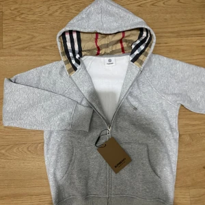Grå hoodie från Burberry med dragkedja - Snygg grå hoodie från Burberry med klassiskt rutigt foder i huvan och dragkedja framtill. Tröjan har broderad Burberry-logga på bröstet, snörning i huvan och känguruficka. Perfekt för dig som vill ha en stilren och lyxig look.