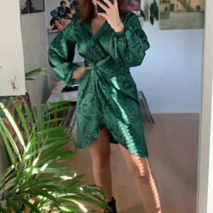 Grön omlottklänning med paisleymönster - Snygg grön kortklänning med svart paisleymönster och omlottdetalj i midjan. Klänningen har långa puffärmar, krage och v-ringad hals. Materialet är glansigt och känns lyxigt.