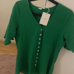 Grön blus med knappar från MQ - Snygg grön blus från Dobber med struktur i tyget och vita knappar framtill. Blusen har korta ärmar och v-ringning, perfekt för dig som vill ha en färgstark och trendig topp. Enkel att matcha med både jeans och kjol.