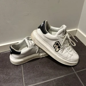 Karl Lagerfeld vita sneakers - Snygga vita sneakers från Karl Lagerfeld med svart hälparti och coolt logomärke på sidan. Skorna har snörning, rund tå och platt sula. Tillverkade i skinn med en chunky design som ger en modern look. Perfekta för dig som vill sticka ut med stil.