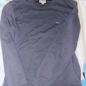 Grå tracksuit från Lacoste M - Snygga grå mjukisbyxox samt sweatshirt från Lacoste i storlek M med tapered fit. Byxorna har elastisk midja med snörning och den klassiska Lacoste-loggan på höften. Perfekta för chill eller träning, tillverkade i mjukt bomullsmaterial för maximal komfort.