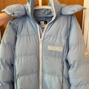 Ljusblå pufferjacka från Hoodrich  - Säljer en ljusblå pufferjacka från Hoodrich i storlek S. Jackan har vit dragkedja, broderad logga på bröstet och stor HOODRICH-text på ryggen. Fodrad och med hög krage, perfekt för kalla dagar.