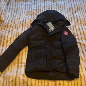 Svart dunjacka från Canada Goose M - Säljer en svart dunjacka från Canada Goose i storlek M. Jackan har huva, dragkedja och klassisk röd logotyp på ärmen. Perfekt för kalla dagar med sin varma och fluffiga känsla. Snygg och stilren design som funkar till det mesta.