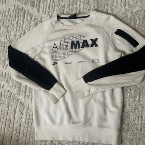 Nike Air Max sweatshirt med svarta detaljer - Nike Air Max sweatshirt i vit med svarta paneler på ärmarna och coolt grafiskt tryck framtill. Tröjan har rund hals, lång ärm och en dragkedjedetalj på ena ärmen. Perfekt för dig som gillar streetwear och sportig stil.