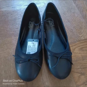 Svarta ballerinaskor med rosett - Stilrena svarta ballerinaskor med rund tå och söt rosettdetalj framtill. Skorna har en platt sula och är tillverkade i syntetmaterial som ger en klassisk look. Perfekta för dig som gillar enkel och tidlös stil.