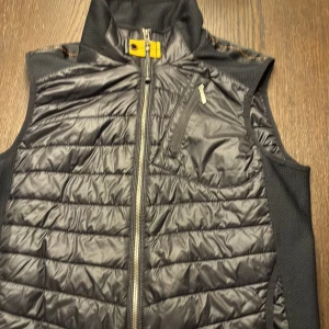 Svart dunväst från Parajumpers - Svart dunväst från Parajumpers med quiltad design och hög krage. Västen har dragkedja framtill, bröstficka med dragkedja och detaljer i orange vid nacken. Snygg och sportig look med diskret logga på ryggen.