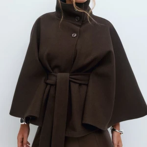 Mörkbrun poncho kappa från Gina tricot - Stilren mörkbrun kappa med hög krage, knappar framtill och brett knytbälte i midjan. Vida ärmar och elegant, minimalistisk design som ger en cool och sofistikerad look. 
