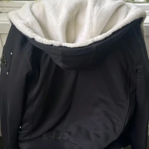 Svart jacka från Moose Knuckles - Säljer en svart jacka från Moose Knuckles i storlek M. Jackan har vit päls i huvan, dragkedja framtill och ikonisk logga på ärmen. Utsidan är slät och insidan har mönstrat foder. Perfekt för kalla dagar.