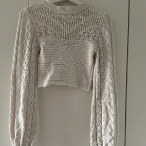 Vit stickad croppad topp Chiquelle - Säljer en vit croppad stickad topp från Chiquelle med långärmade ärmar och snyggt virkat mönster. Toppen har snörning vid ärmsluten och en högre hals. Perfekt för dig som gillar boho-vibe och detaljerad stickning.