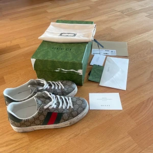 Gucci Ace skor - Gucci Ace sneakers i canvas med klassiskt GG-monogram, gröna och röda ränder på sidorna och vita snören. Skorna har vit gummisula med mönster och vita detaljer vid hälen. Kommer med originalkartong, dustbag och tillbehör. Perfekta för dig som vill sticka ut med lyxig streetstyle. Skriv för mer info (ÄKTA KVITTO FINNS)