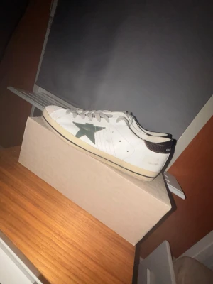 Golden Goose Superstar sneakers vit/grön - Golden Goose Superstar sneakers i vitt skinn med grön stjärna på sidan och grå snörning med logotyp. Bakkappa i mörkbrunt med metallicfinish. Platt sula och rund tå. Snygg retrostil med beige detaljer på sulan och tungan.