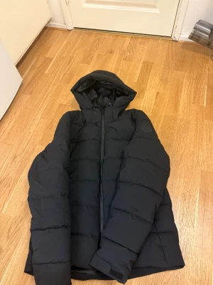 Svart pufferjacka med huva - Säljer en helt ny svart jacka i modern, stilren design. Jackan är aldrig använd och är i perfekt skick. Den är vadderad och mycket varm, perfekt för kalla höst- och vinterdagar. 