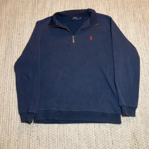 Marinblå half zip tröja Polo Ralph Lauren - Snygg marinblå långärmad tröja från Polo Ralph Lauren med röd broderad logga på bröstet. Tröjan har en half zip-dragkedja vid halsen och är gjord i mjuk bomull. Perfekt för chill dagar och enkel att matcha med jeans eller chinos.