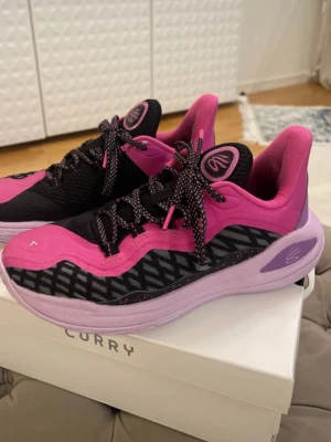 Svarta och rosa Curry basketskor - Säljer ett par snygga Curry sneakers i svart och rosa med mönstrade detaljer på sidorna. Skorna har snörning med svarta och vita skosnören, mesh-överdel för bra ventilation och en mjuk, ljuslila sula. Perfekta för dig som gillar sportig stil och färg.