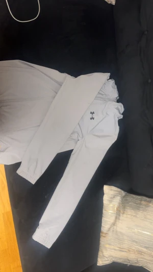 Grå  vindjacka från Under Armour - Säljer en fräsch grå vindjacka från Under Armour med snyggt tryck på ryggen och logga på bröstet. Jackan har dragkedja framtill, snörning vid kragen och långa ärmar med resår vid mudden. Perfekt för träning eller chill dagar ute.