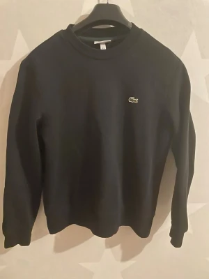 Svart sweatshirt från Lacoste - Klassisk svart sweatshirt från Lacoste i bomull med rund hals och broderad grön krokodil på bröstet. Tröjan har lång ärm och ribbade muddar för en clean och stilren look. Perfekt till höst och vinter när du vill ha något enkelt men snyggt.